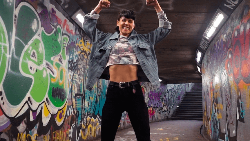Image: Sorelle in a tunnel of identity graffiti, smiling and posing with her arms flexed above her head